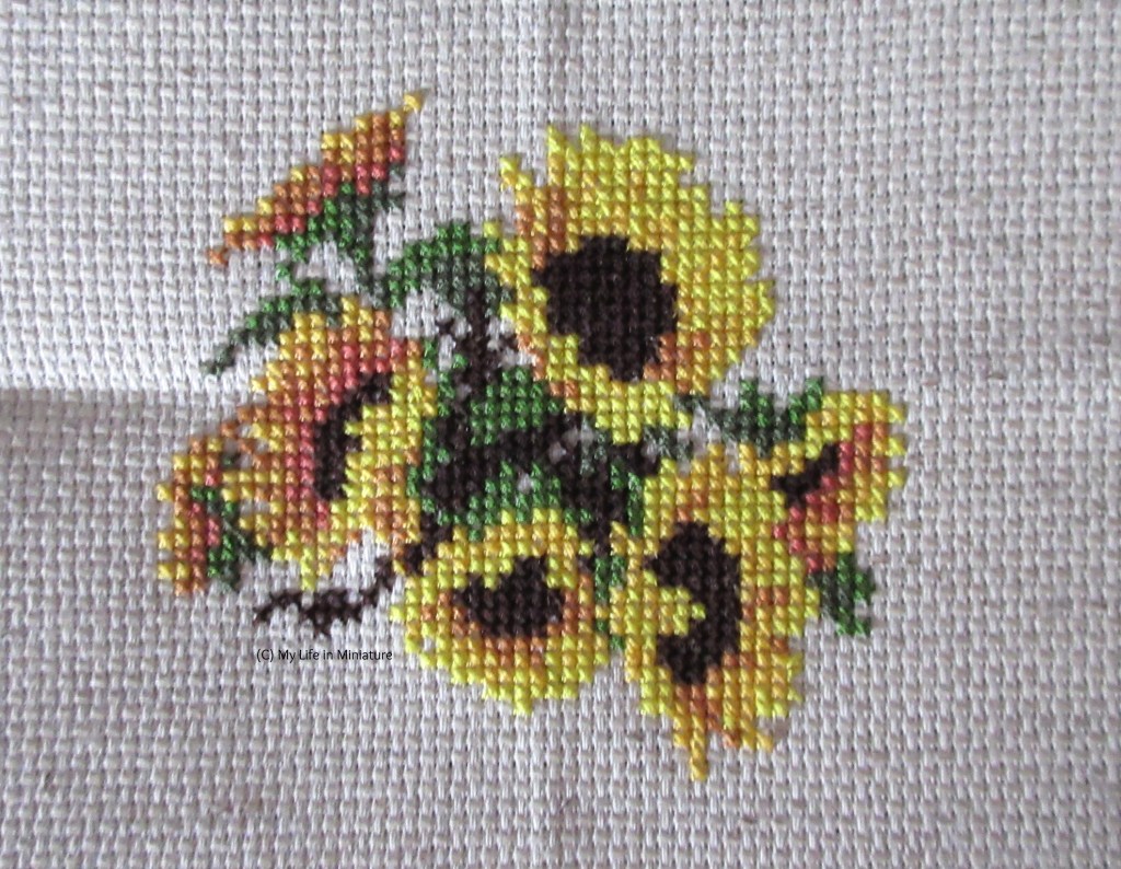 Seven sunflowers, with leaves and stems behind them, are cross-stitched onto cream fabric. 