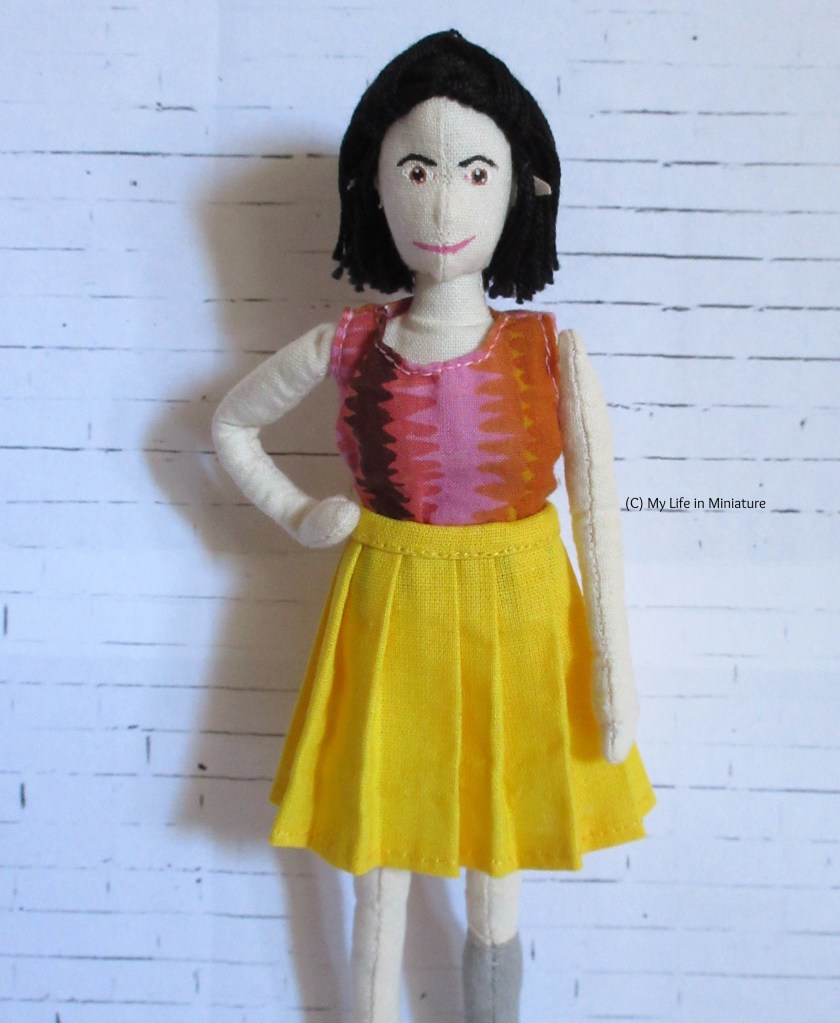 Tiffany stands in front of a white brick background facing the camera. She wears a yellow pleated knee-length skirt, and  a pink-and-orange top. She has a hand on her hip.