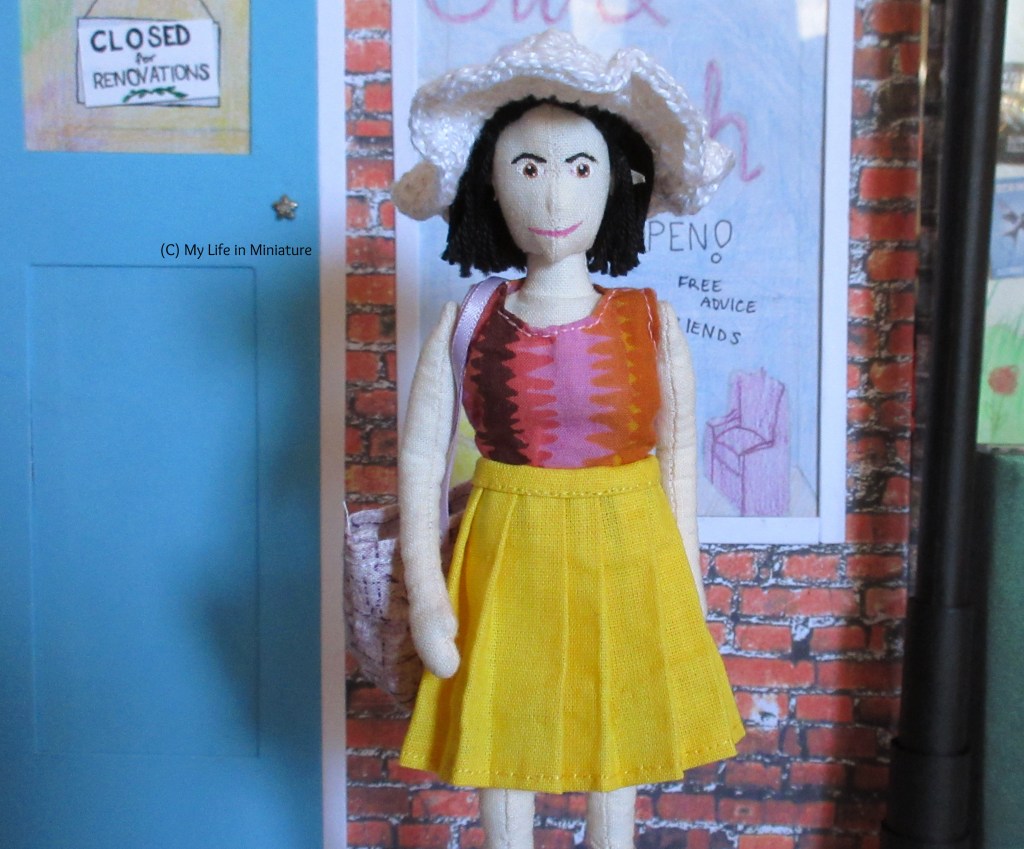 Tiffany stands outside Needle & Thread. She wears the yellow pleated skirt, pink-and-orange top, and a white crocheted sun hat. She carries a pale purple tote bag, and looks straight at the camera.