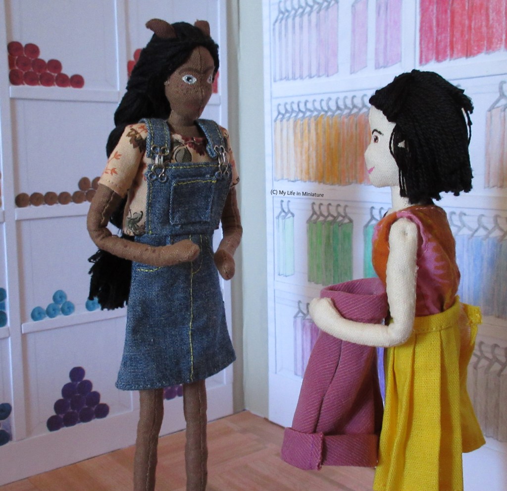 Tiffany holds the completed pair of corduroy shorts to her waist, looking at Petra for her reaction. Petra is clapping her hands together, looking at the shorts. They are in the fat quarters/yarn shelves corner of Needle & Thread.