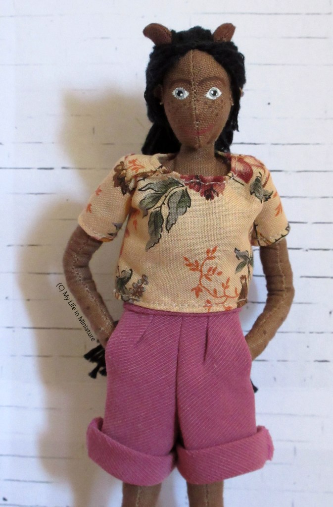 Petra looks at the camera, hands in pockets of her dusky pink corduroy shorts. There are two pleats in the front of the pants, and the bottom hems are rolled up. She wears her cream floral t-shirt, and stands in front of a white brick background.
