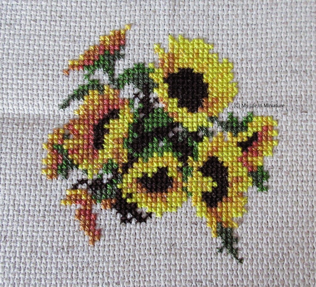 Seven complete sunflowers and the beginnings of an eighth are cross-stitched onto cream fabric, with green leaves around and in-between them. 