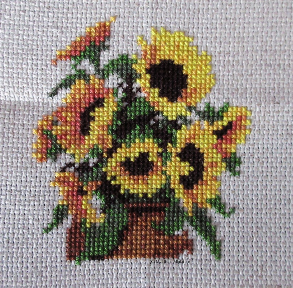 Eight sunflowers, their leaves and stems, and a brown basket beneath them are cross-stitched onto cream fabric. 