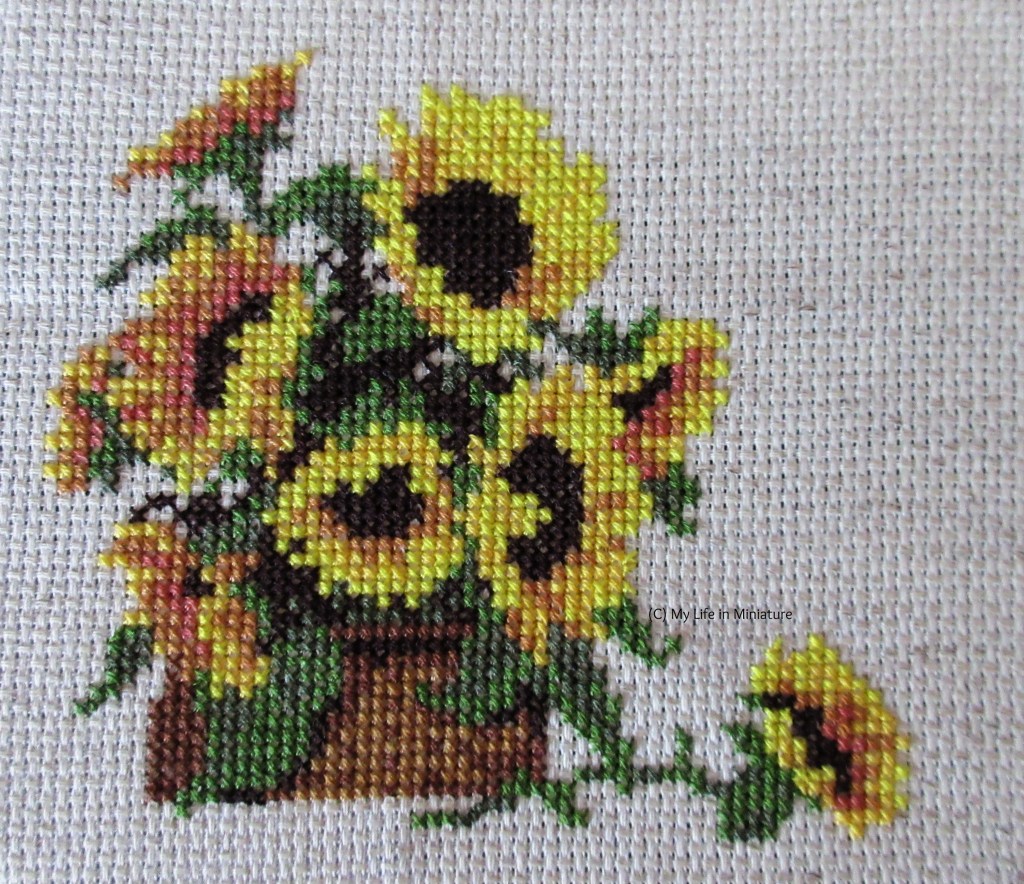 Eight sunflowers in a basket are cross-stitched onto cream fabric. In front of the basket is a ninth sunflower (with stem and leaves) that has fallen out of the basket, cross-stitched.