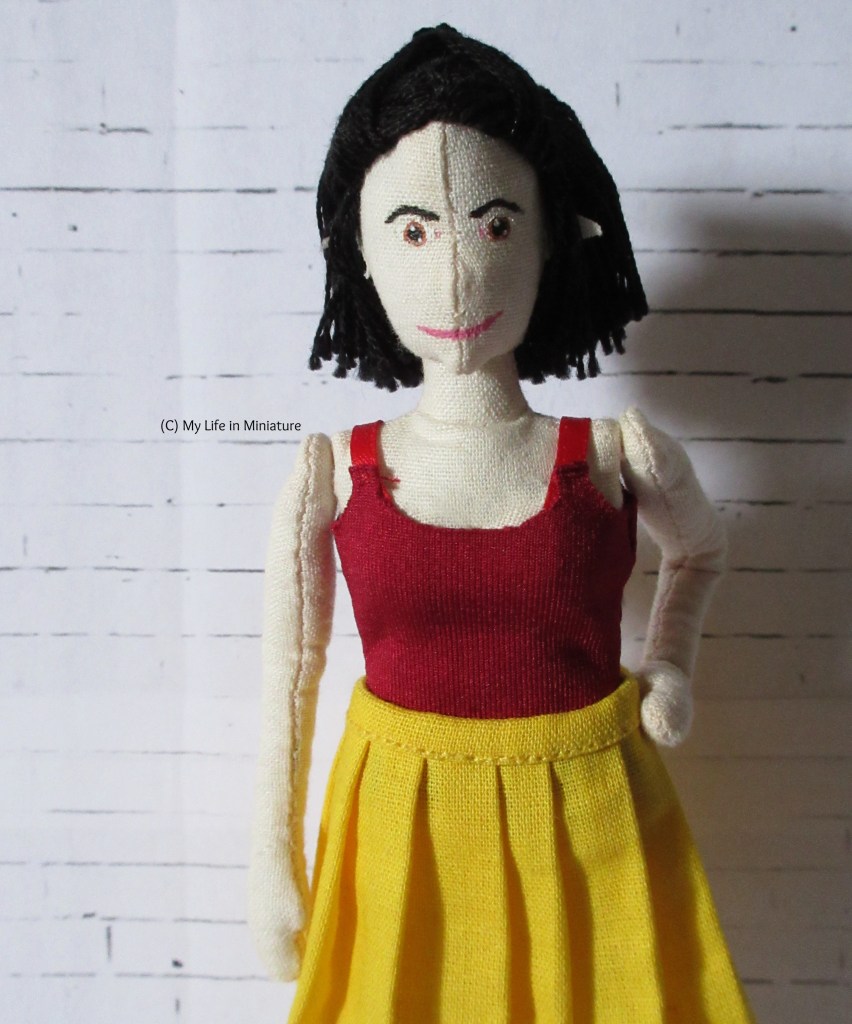 Tiffany wears a dark red jersey scoop-necked singlet top, which has red ribbon for straps. She also wears the yellow pleated skirt, and stands with a hand on hip in front of a white brick background.