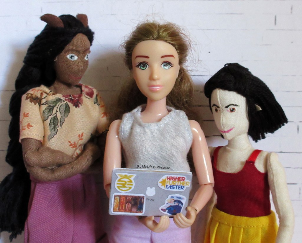 Sarah, Tiffany and Petra sand around Sarah's open laptop. Sarah is in the centre, holding the laptop and looking at the camera. Tiffany and Petra are looking over Sarah's shoulders at the laptop. 