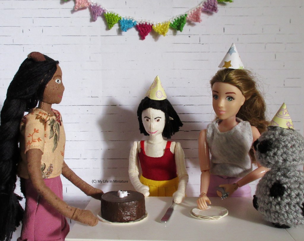 Sarah, Tiffany and Petra stand around a white table having a party. Bunting is on the white brick background. Chip, Sarah's android, is on the table on the right, in a party hat and facing left. Petra is on the left, hands on the table and looking at Sarah and Tiffany. Tiffany is eagerly reaching for the large knife to cut the cake with, wearing a party hat. Sarah is next to Chip on the right, preparing the plates and forks for the pieces of cake. She and Chip are wearing party hats.