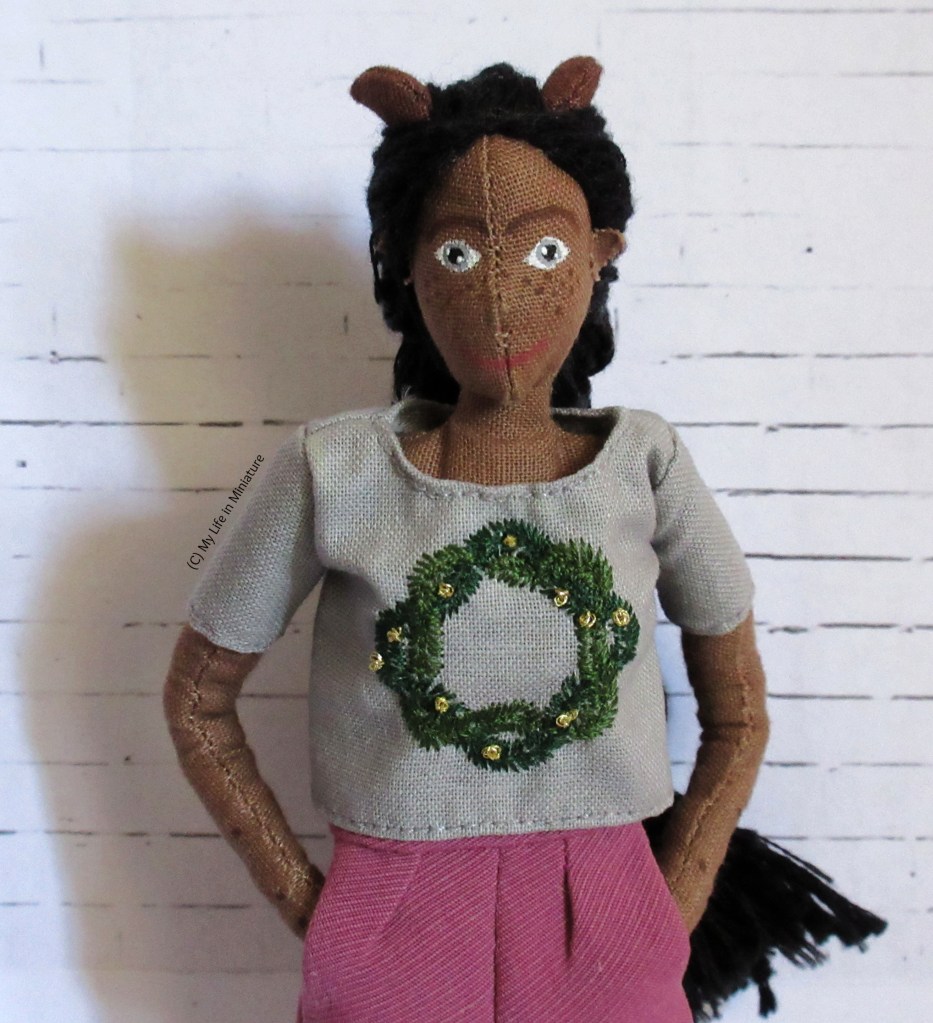 Petra stands in front of a white brick background, hands in pockets. She looks at the camera and is wearing a grey t-shirt with a green embroidered wreath on the front. 