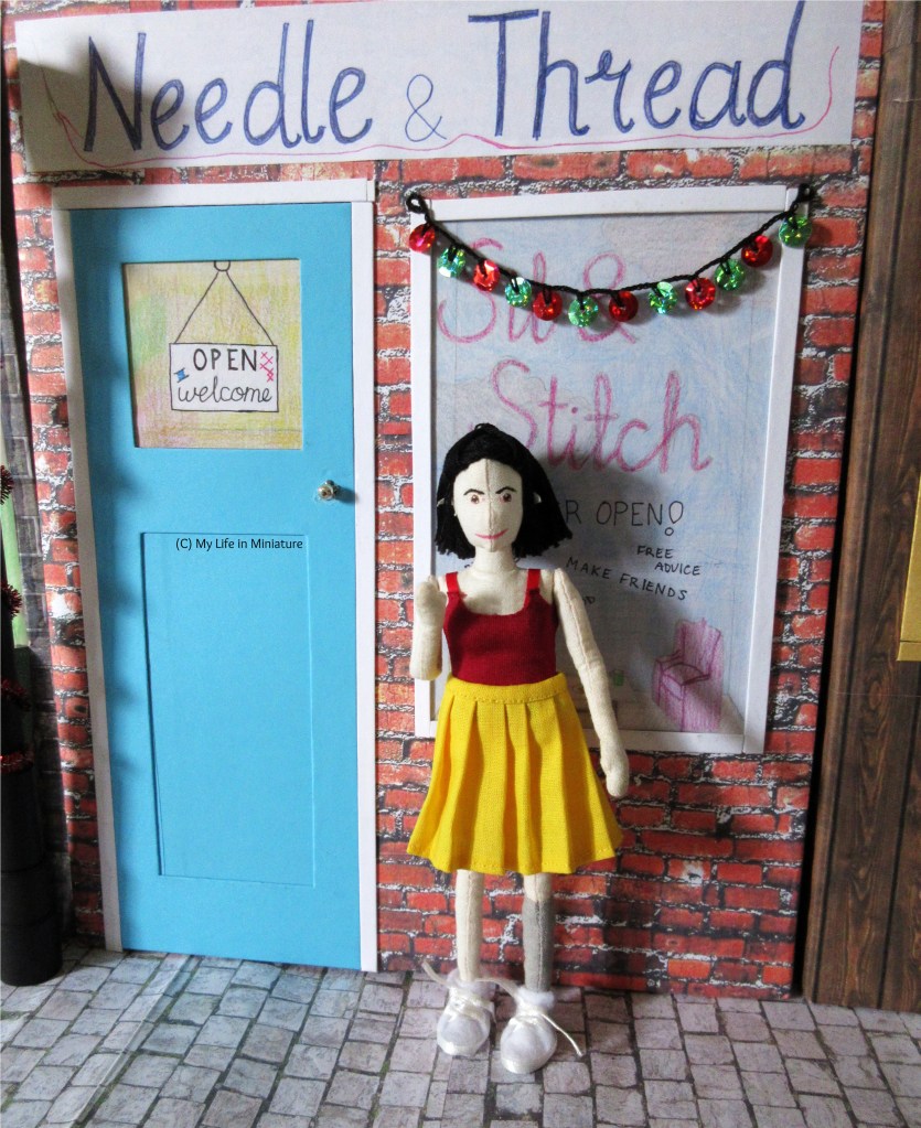 Tiffany stands outside Needle & Thread, waving at the camera. Needle & Thread's door has a sign in the window saying 'Open; welcome', and the front window is decorated for Christmas. 