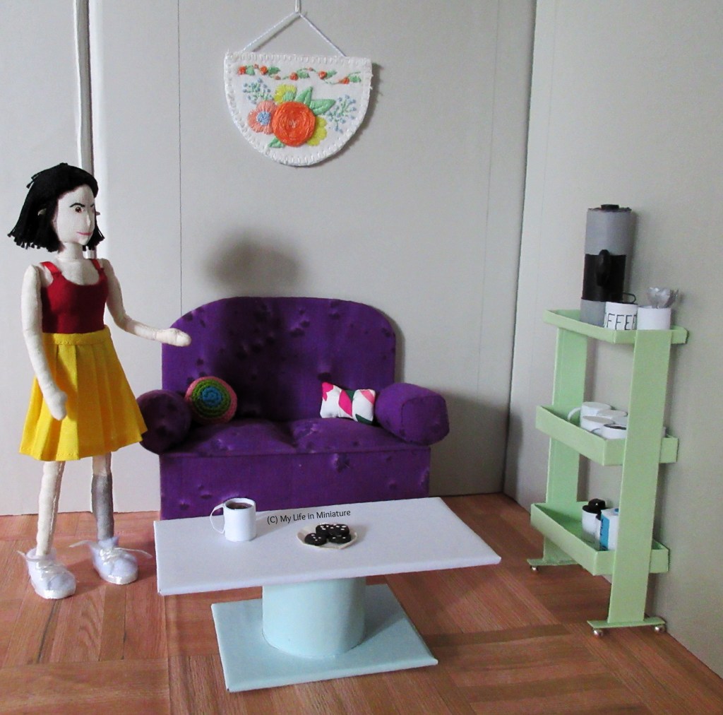 Tiffany stands to the left of the sit and stitch area, gesturing to the space. There are biscuits on the table and an embroidery hangs on the wall behind the couch.