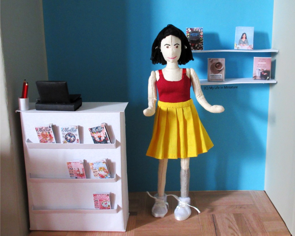 Tiffany stands in the middle of the frame. To her left is the counter and to her right are two shelves attached to the blue wall behind her. There are four craft books on the shelves, displayed on stands.