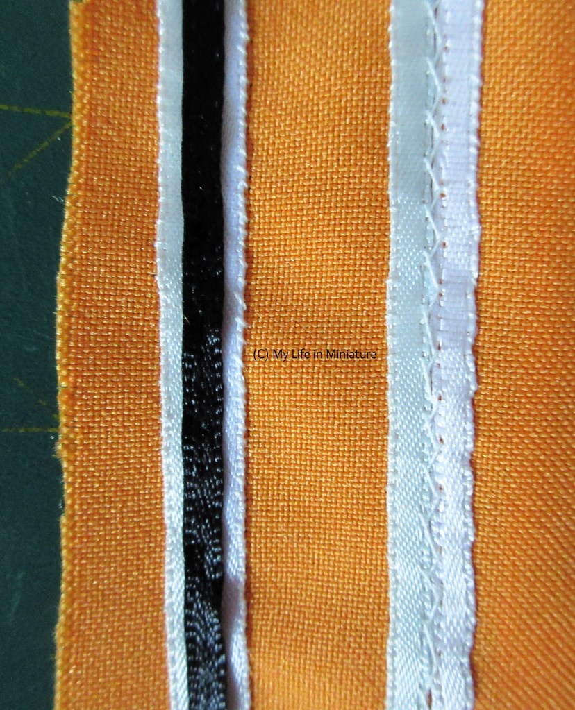 Overhead shot of two stripes in progress. The stripe on the right has a strip of pale blue ribbon sewn next to a strip of white ribbon. They are herringbone stitched together down the centre. The stripe on the left is complete, with the black ribbon back-stitched down the centre. 