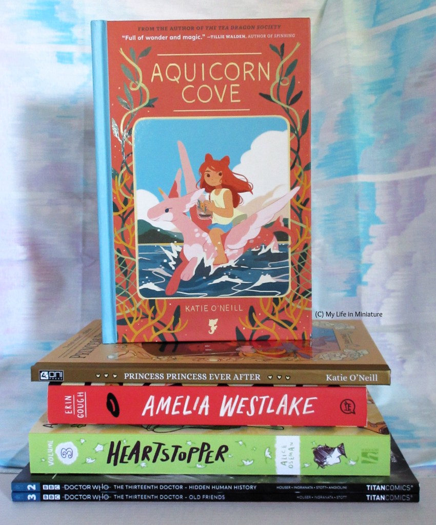 'Aquicorn Cove', 'Princess Princess Ever After', 'Amelia Westlake', Heartstopper vol. 3 and the two Thirteenth Doctor comics sit in a pile. 'Aquicorn Cove' is standing on top so the cover is visible. 