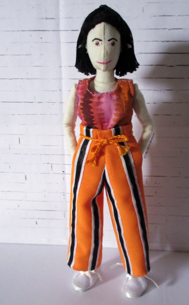Tiffany stands in front of a white brick background, looking at the camera. She wears a t-shirt and a pair of bright orange pants. The pants have vertical black stripes which are sandwiched in-between pale blue and white stripes. They also have an orange plaited cord tied around the waist and pockets. 