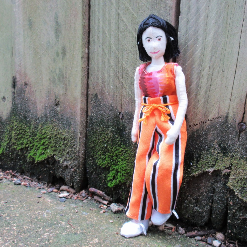 Tiffany leans against a wooden fence, one foot up against the wood, wearing the orange striped pants. There is green moss along the base of the fence, and she is looking at the camera. 