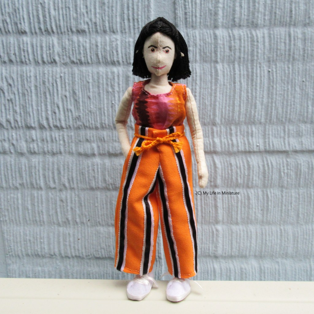 Tiffany stands against a pale blue textured wall, looking at the camera. She wears the orange striped pants, one hand in a pocket. 