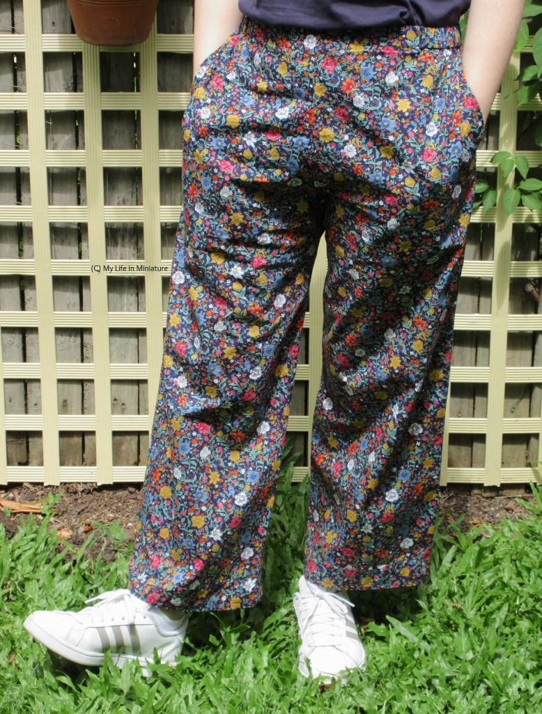 The author wears the floral pants, hands in pockets, and white shoes. She is outside, standing against a fence. 