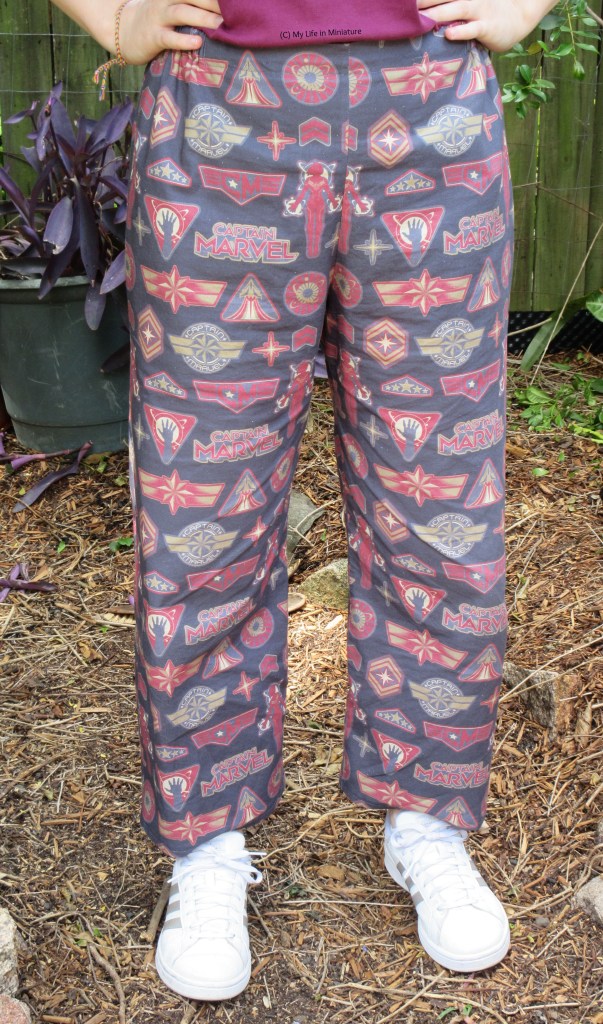 The author wears loose, elastic-waisted pants printed with a Captain Marvel-inspired design with white shoes. She stands outside on a garden pathway, hands on hips.