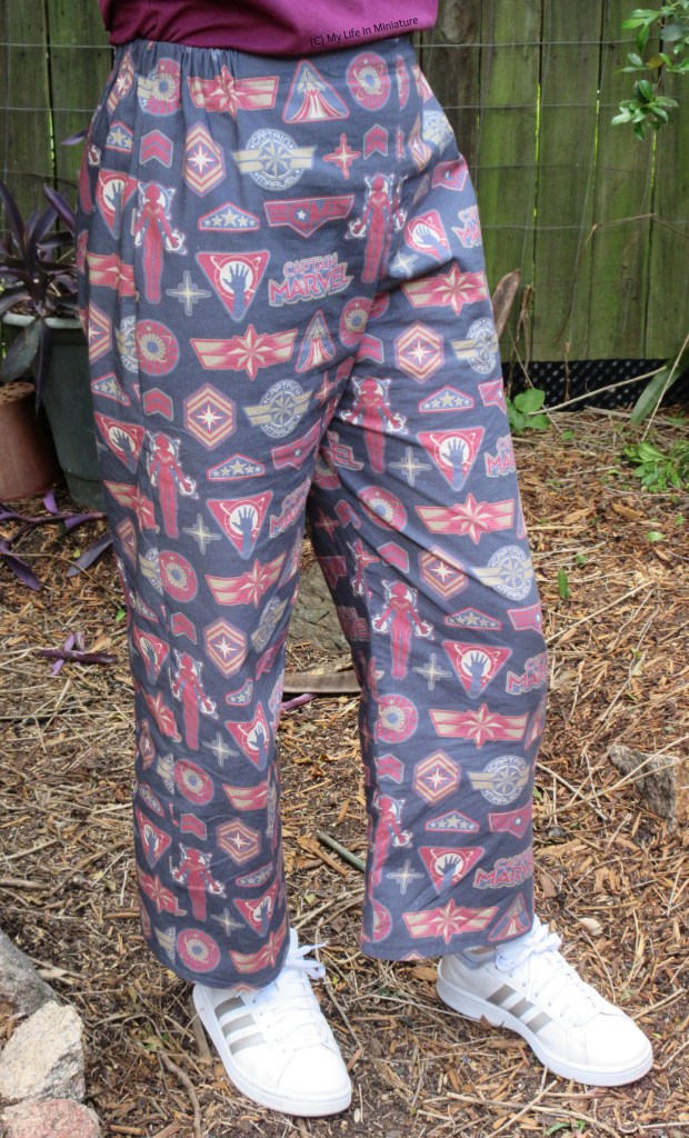 The author wears loose, elastic-waisted pants printed with a Captain Marvel-inspired design with white shoes. She stands outside on a garden pathway, one knee bent slightly.