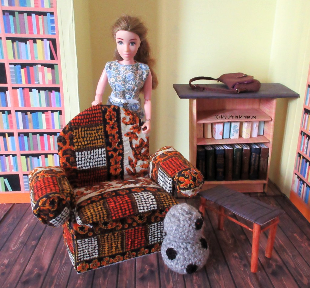 Sarah stands inside The Palace Library. She has her hands on the back of a brightly patterned armchair, looking down at it. Chip is in front of the armchair, looking at a small side table. The counter is in the background with Sarah's bag on it.