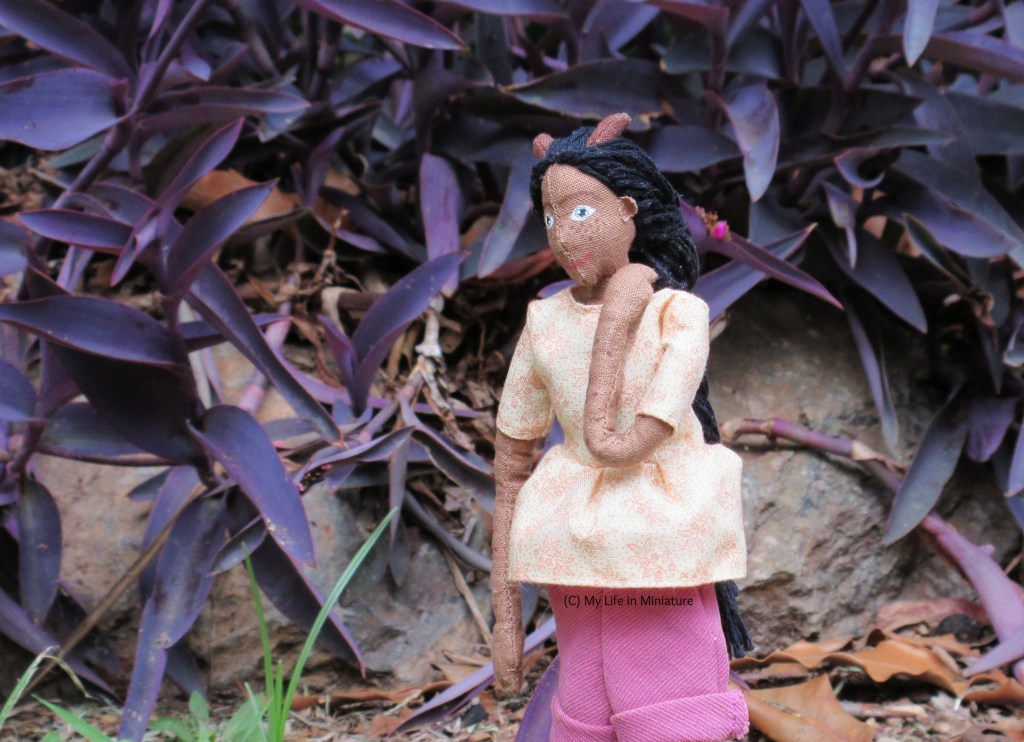 Petra stands outside, in front of a rock-bordered garden bed filled with a purple-leafed plant. She looks to the bottom-left, and has one hand on her shoulder. She wears the gathered cream t-shirt and pink corduroy shorts.