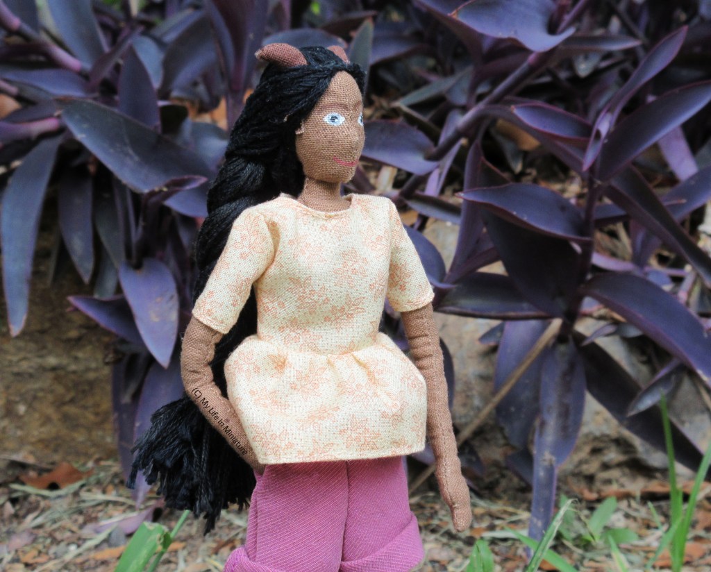 Petra stands outside, in front of a rock-bordered garden bed filled with a purple-leafed plant. She looks to the right, with one hand in her pocket. She wears the cream gathered t-shirt and pink corduroy shorts. 