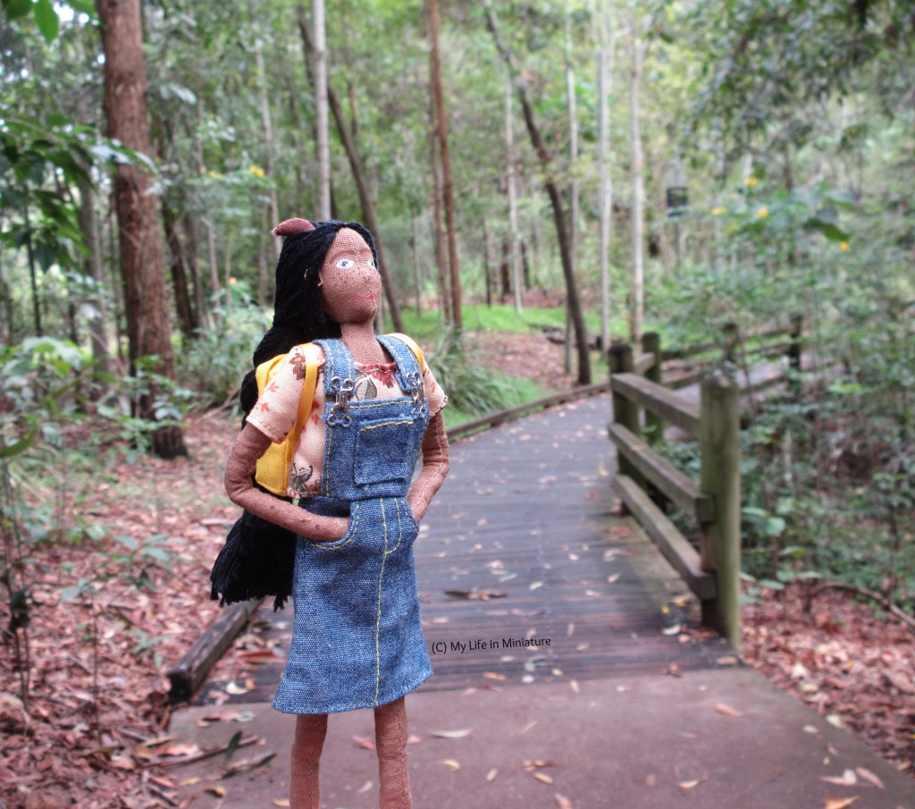 Petra stands on a path that curves away in the distance into a small forest. She looks up beyond the frame at the canopy. She wears her denim pinafore and a cream floral t-shirt and has her yellow backpack on.  