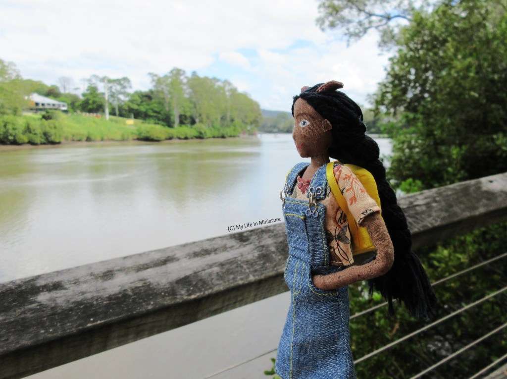 Petra stands at the railing of a boardwalk, hands in pockets. She looks out over the railing at a wide river. The other back is visible, and the water is flat.