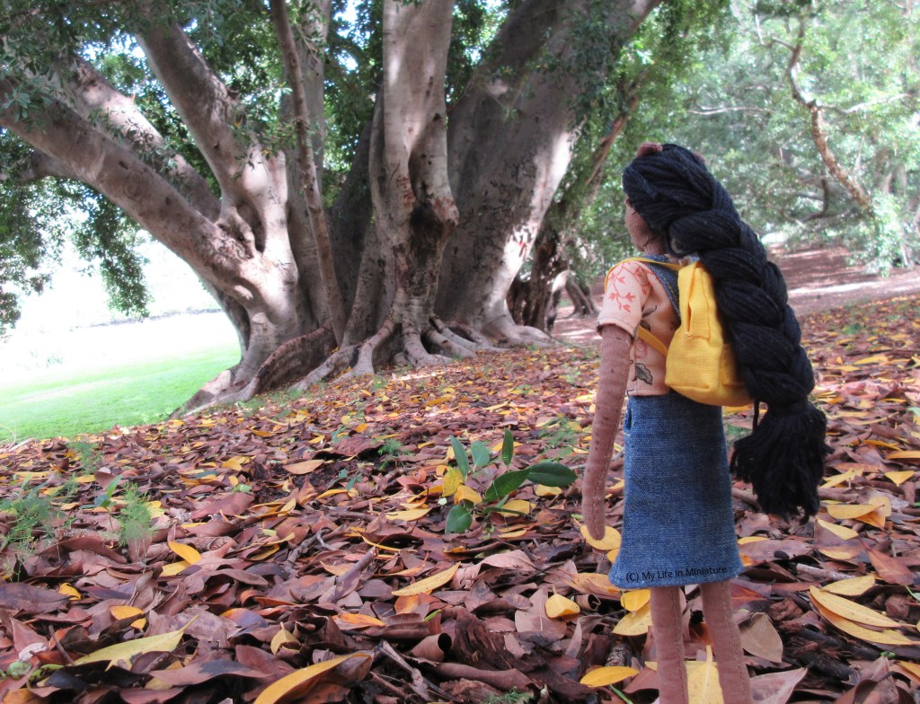Petra faces away from the camera, looking at an enormous tree in the background. It slopes away from her slightly, and on the ground is a thick layer of brown and yellow leaves.