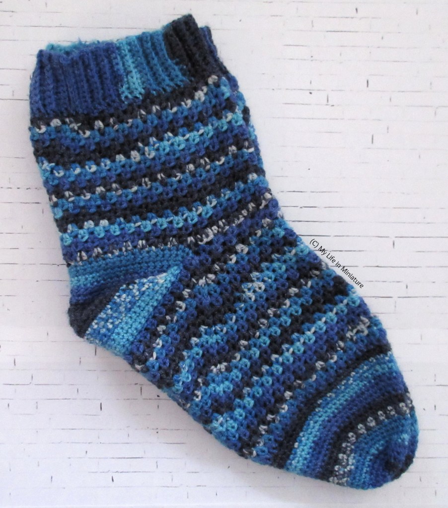A pair of crocheted socks is laid flat on a white brick background. The socks are various shades of blue, with sections of speckled colour. The leg and foot sections are in a granny square-esque stitch, while the heel and toe are solid double crochet.