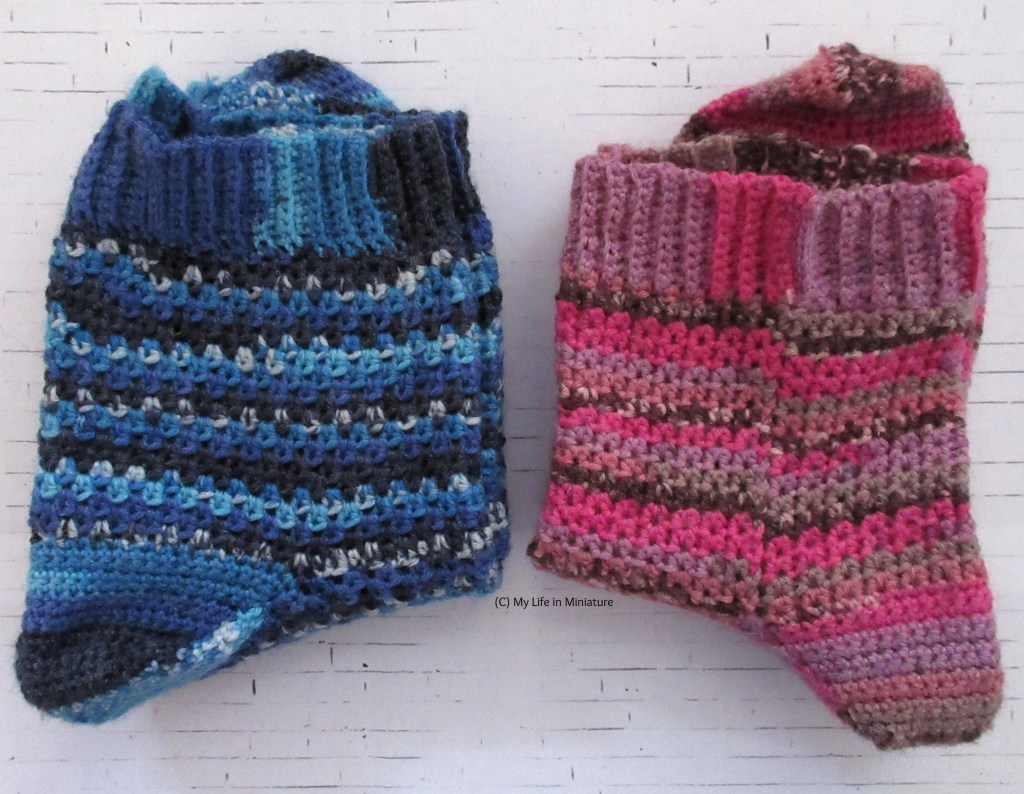 Two pairs of crocheted socks sit folded on a white brick background. The pair on the left are various shades of blue, with sections of speckled colour. The pair on the right are shades of pink, pale purple, grey, and speckled brown.