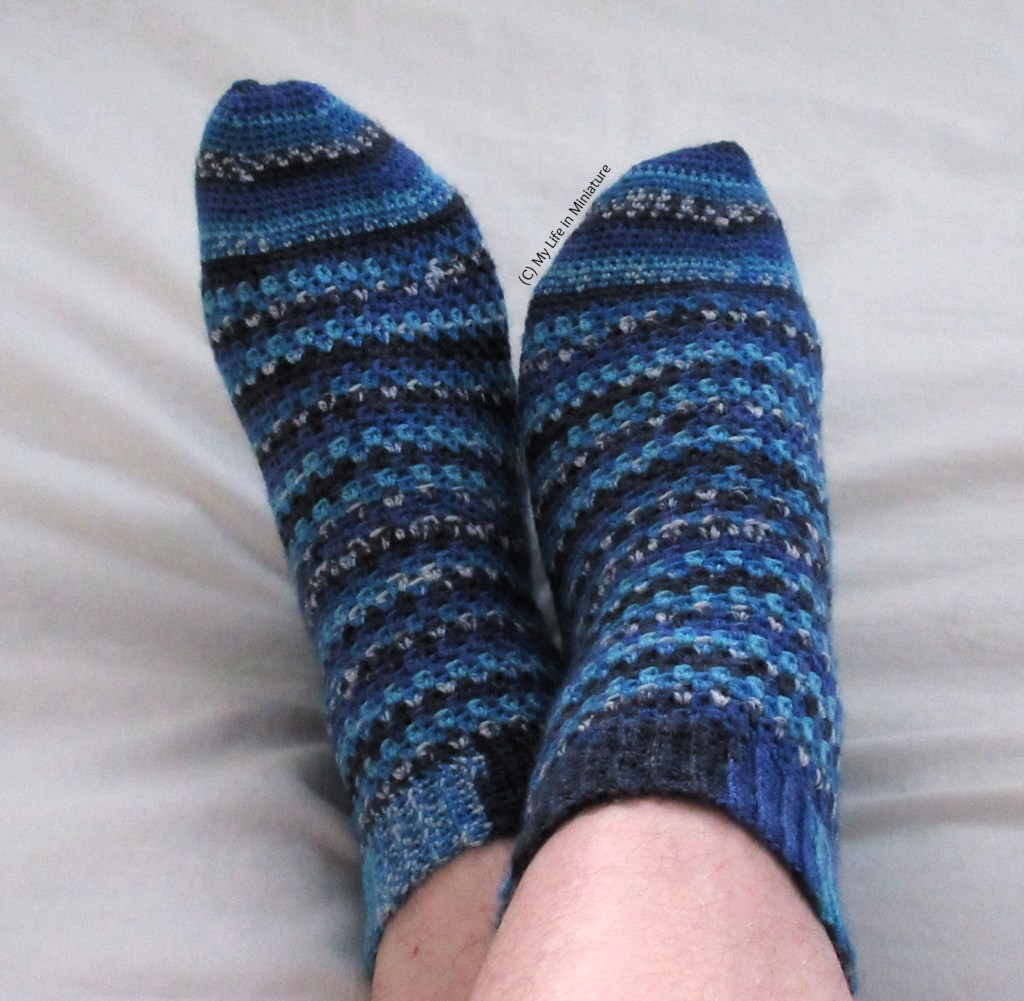 The author wears a pair of crocheted socks on a pale grey fabric background. The socks are various shades of blue, with sections of speckled colour. Her feet are crossed over each other.