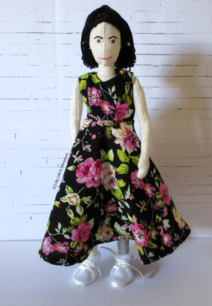 Tiffany stands in front of a white brick background. She wears an ankle-length sleeveless dress with a circular skirt and V-neck. It is black with pink and orange flowers on, and green leaves.