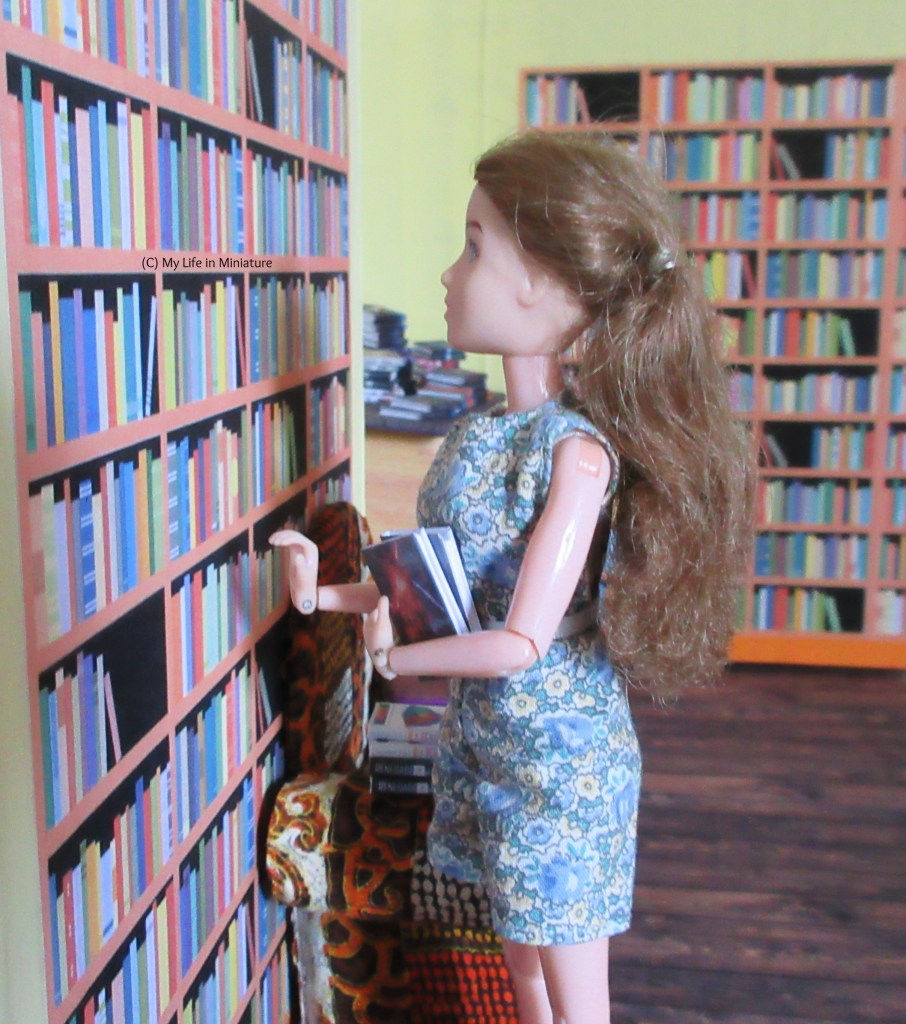 Sarah stands at a bookshelf in The Palace Library, with an armful of books. She looks up at the shelves. Behind her is an armchair, and the front counter, still covered in books.