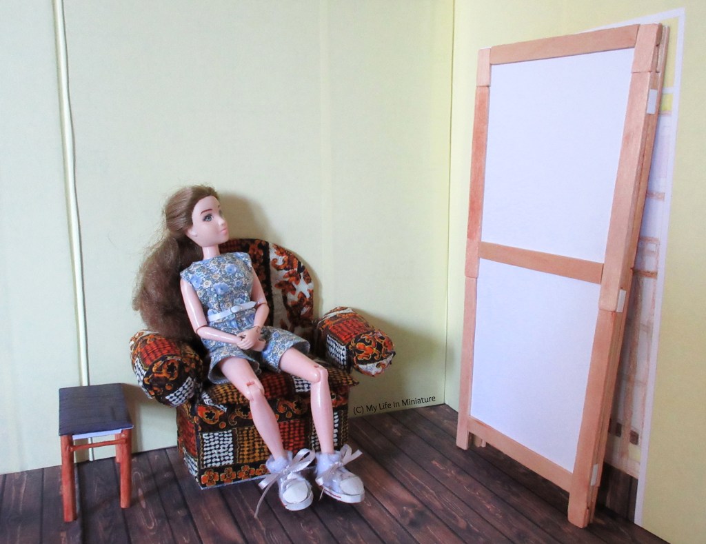 The folded-in-half room divider is standing in front of the doorway of one of The Palace Library's upper rooms. Sarah sits in the red multicoloured armchair against the other wall, looking at the divider. A small side table is beside the armchair.