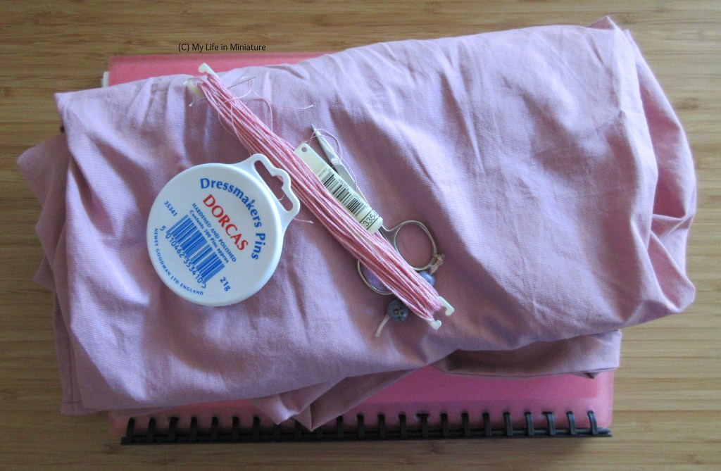 A pile of dusky pink fabric sits on a wood tabletop. On the fabric is a pin tin, a skein of matching embroidery thread, and a pair of embroidery scissors.
