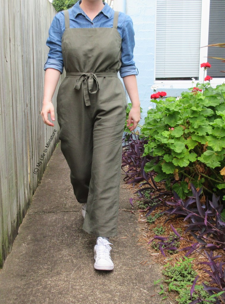The author walks towards the camera, wearing the dungarees over a chambray shirt with white shoes. Her arms are at her sides, and she is on a pat next to a garden bed.