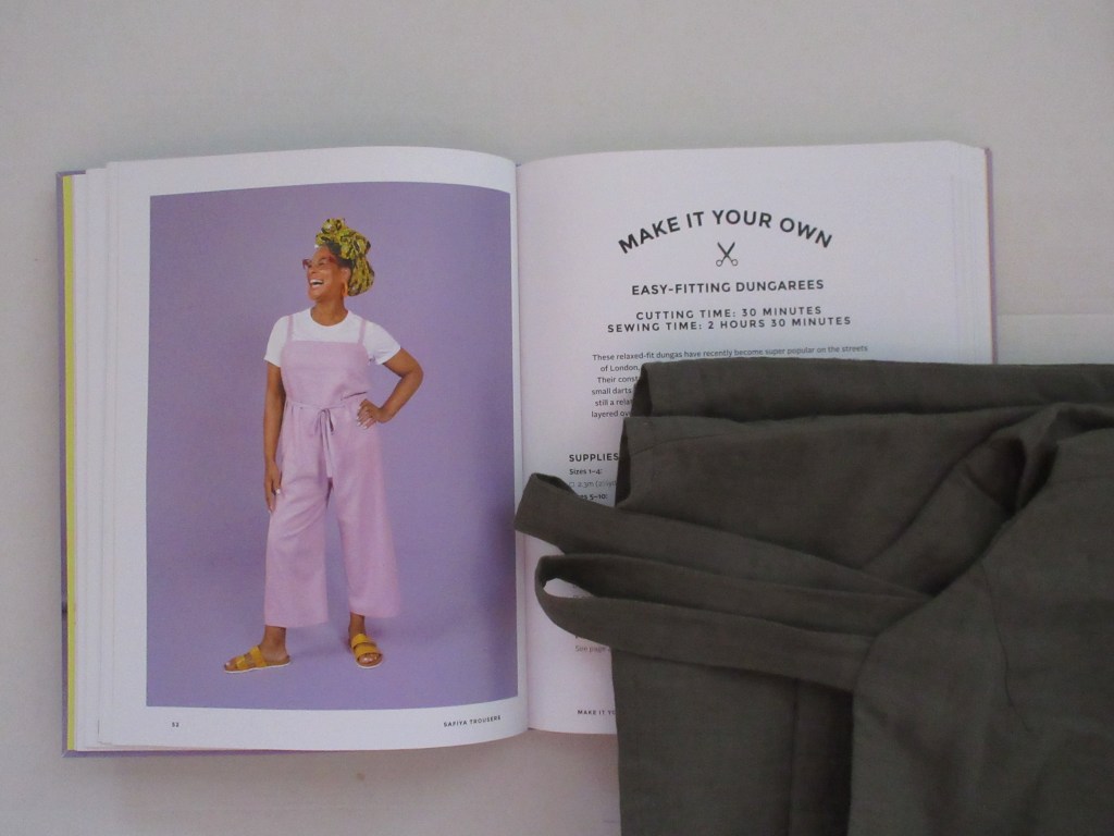 The dungarees lie folded next to an open book on a white background. The book is open to a double page with an image of a woman wearing purple dungarees and the first page of instructions for making the dungarees.