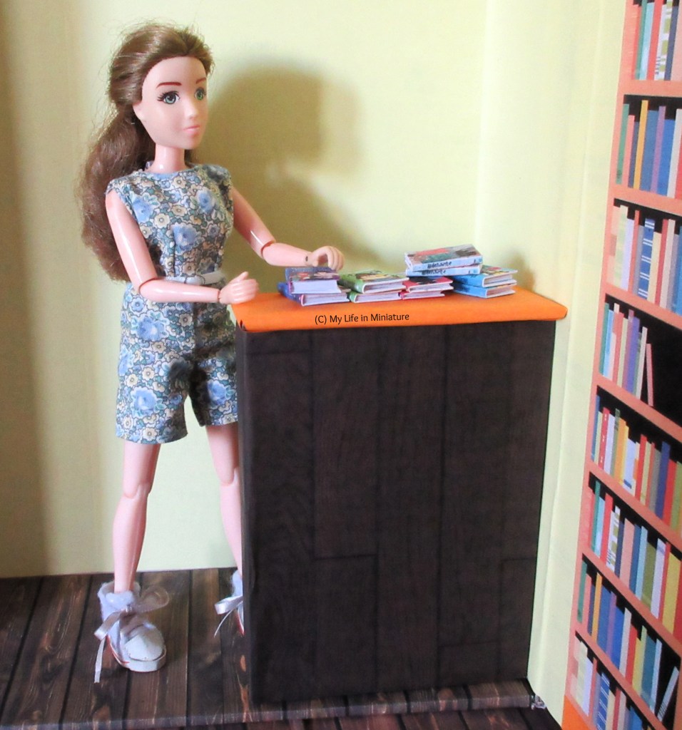 Sarah goes to grab a book from the small pile of novels that is on the new counter at The Palace Library. The counter is dark wood and has a bright orange top. 