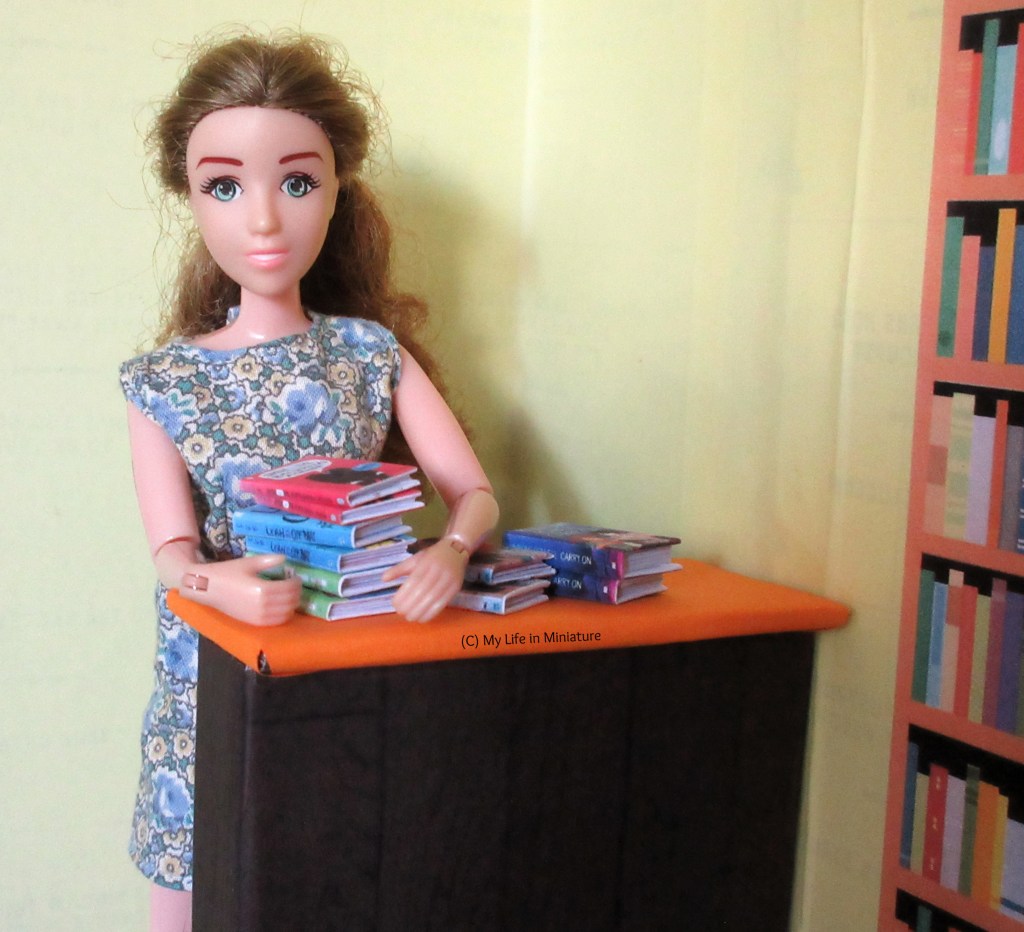 Sarah gathers three of the books from the counter into her arms, ready to shelve. She smiles at the camera. 