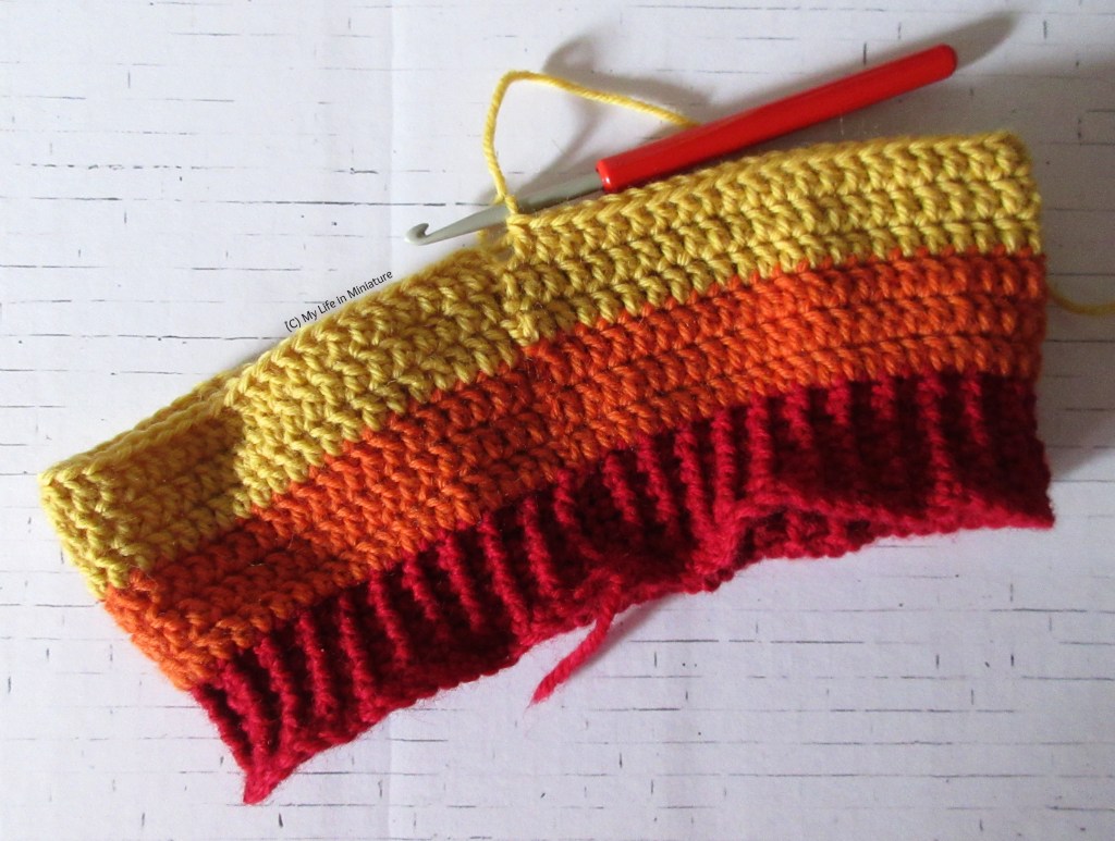 A half-finished crochet beanie sits on a white brick background. The ribbing is red, and the six rows above it are in two stripes; one orange, one yellow. 