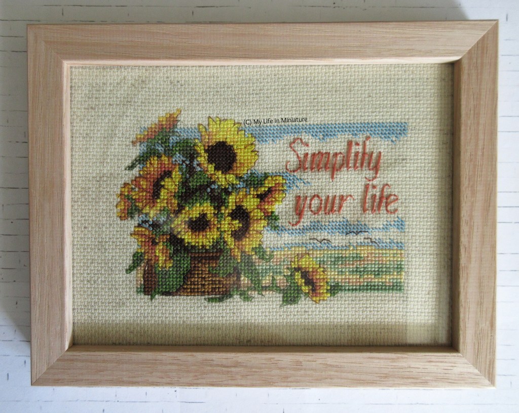 A light wood frame, with glass, sits on a white brick background. In it is a cross-stitching of nine sunflowers in a basket against a field and sky. Text to the right of the sunflowers says 'Simplify your life'. 