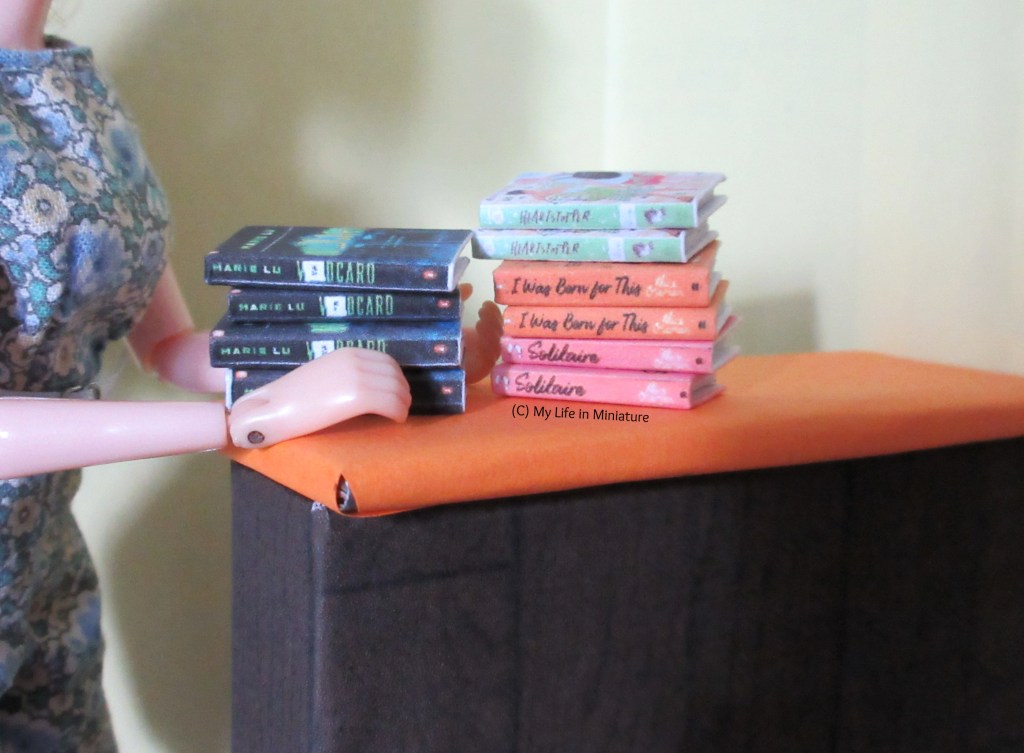 Ten books sit stacked in two piles on the orange countertop in The Palace Library. Sarah's hands are visible around one of the stacks. 