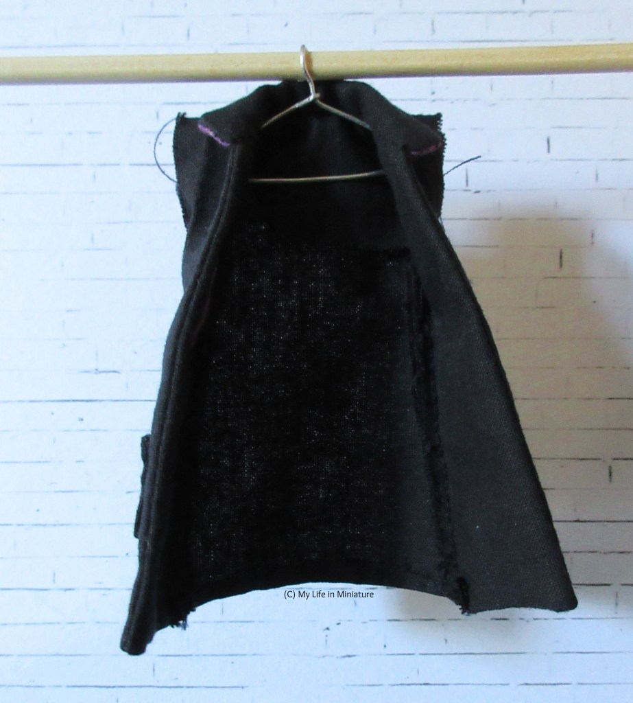 A black coat, unlined and sleeveless, hangs on a coat-hanger from a wooden dowel. 