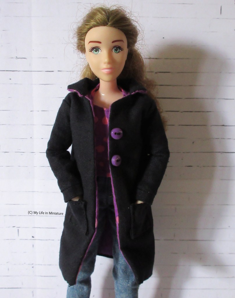 Sarah wears a black, knee-length coat over a t-shirt and a pair of jeans. The coat has a collar, two purple buttons on the front, and pockets, which Sarah's hands are in. She smiles at the camera.