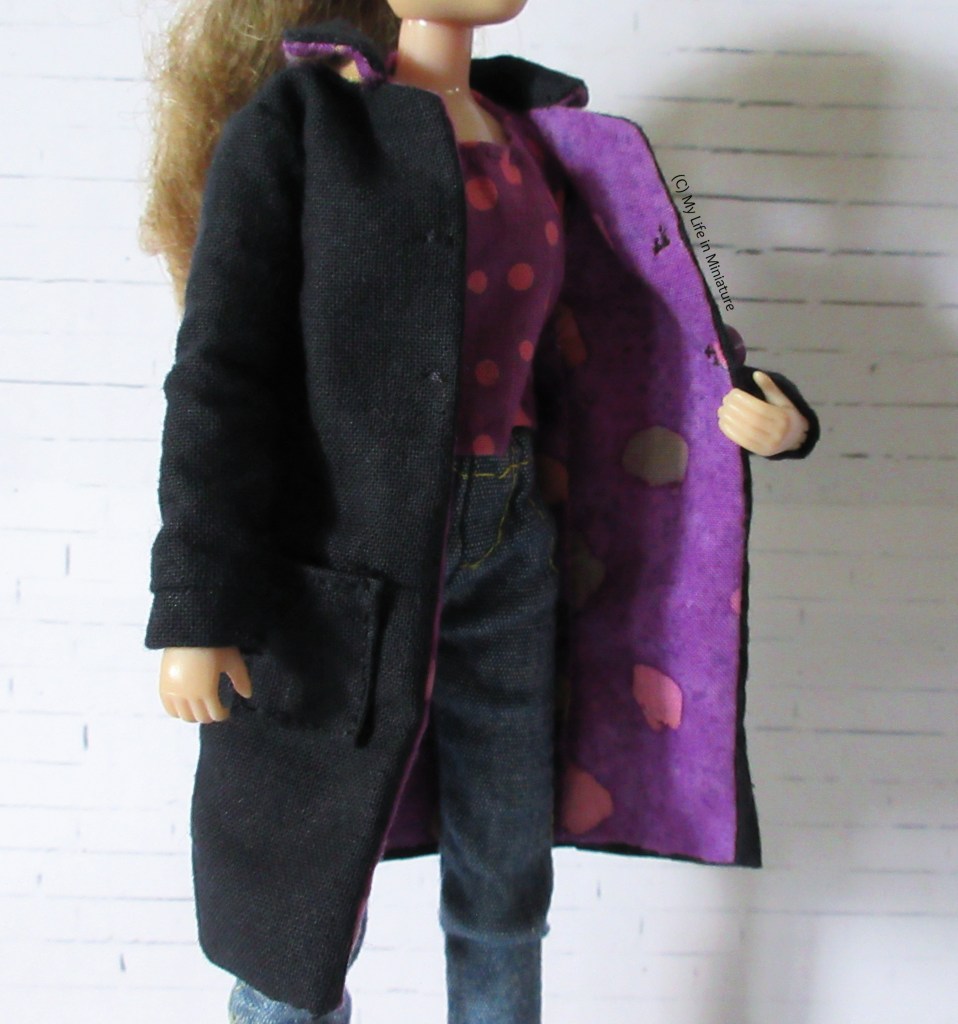 Sarah holds the coat open to show the purple lining. Two stitches on the opposite front panel to the buttons are visible, acting as fake buttonholes. 
