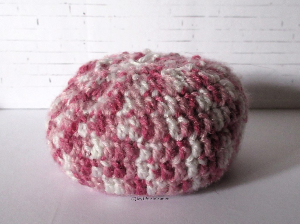 A miniature crocheted pouf sits against a white brick background. It is white and various shades of pink with a flat base.