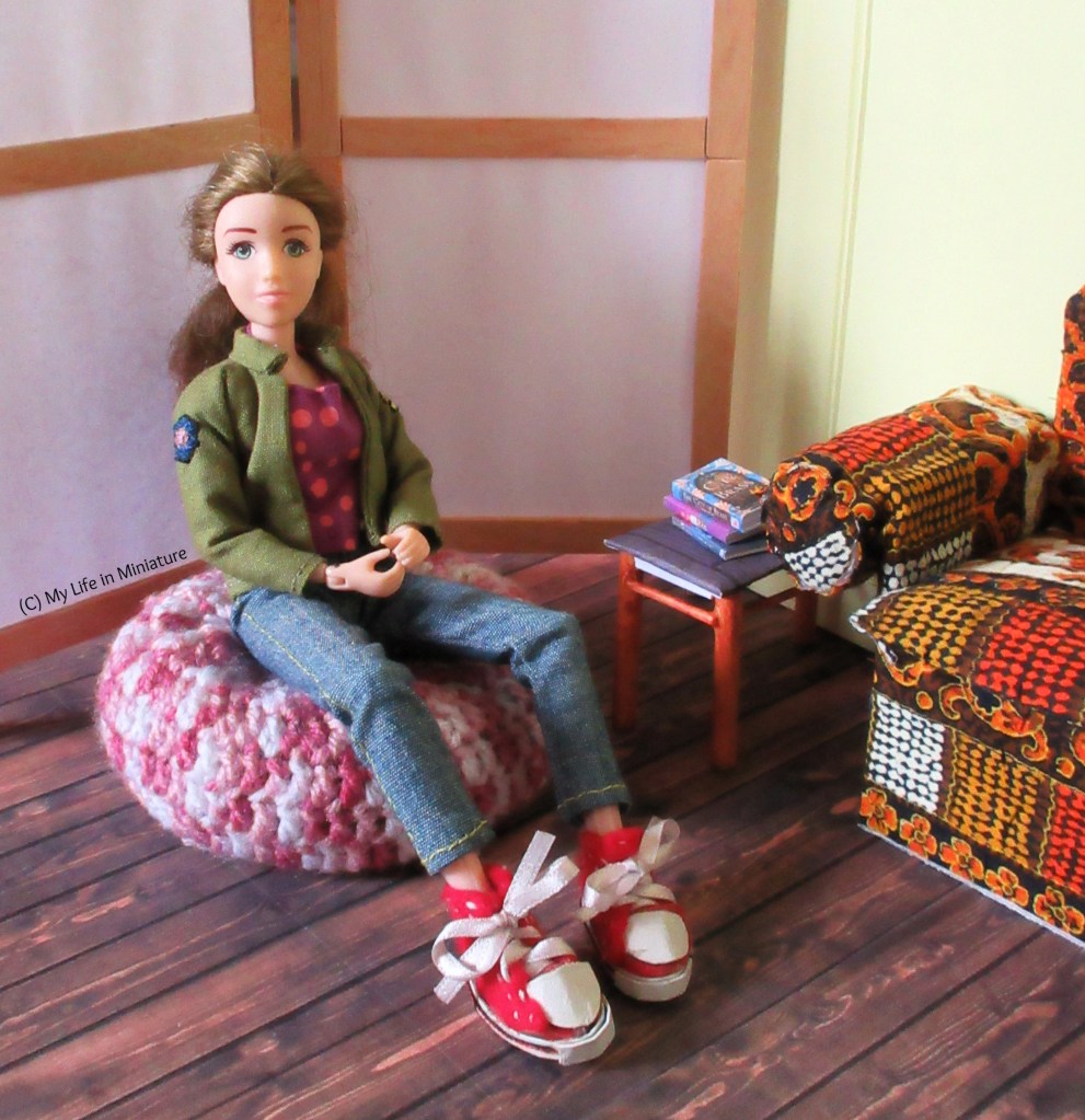 Sarah sits on the beanbag, smiling at the camera. The armchair is in the right of frame, and the side-table with its stack of books is visible.