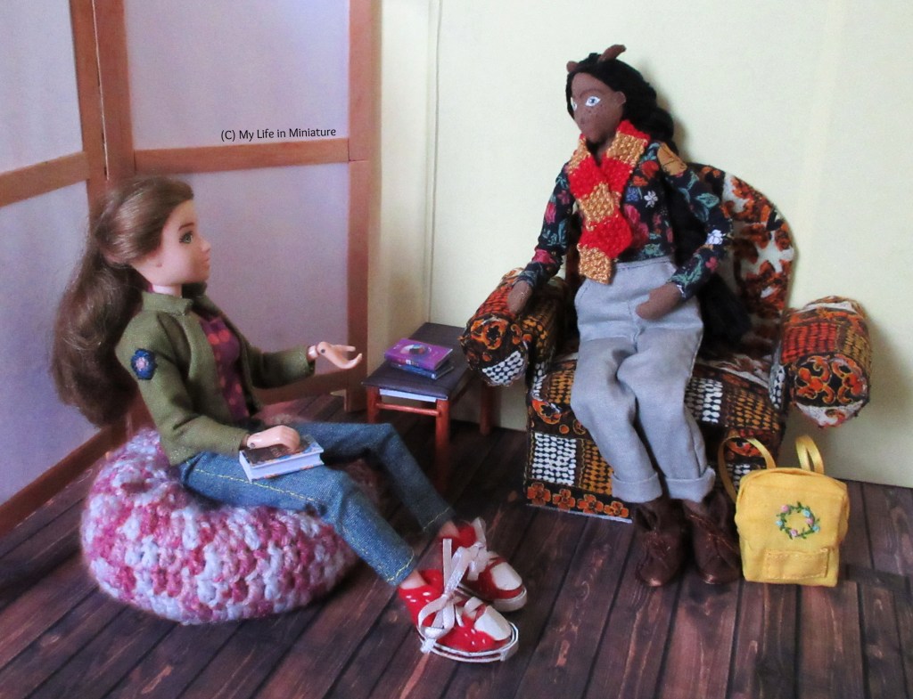 Sarah sits on the beanbag, a book in her lap and gesturing in conversation with Petra. Petra sits on the armchair, legs tucked under, listening to Sarah. Petra's backpack sits at her feet.