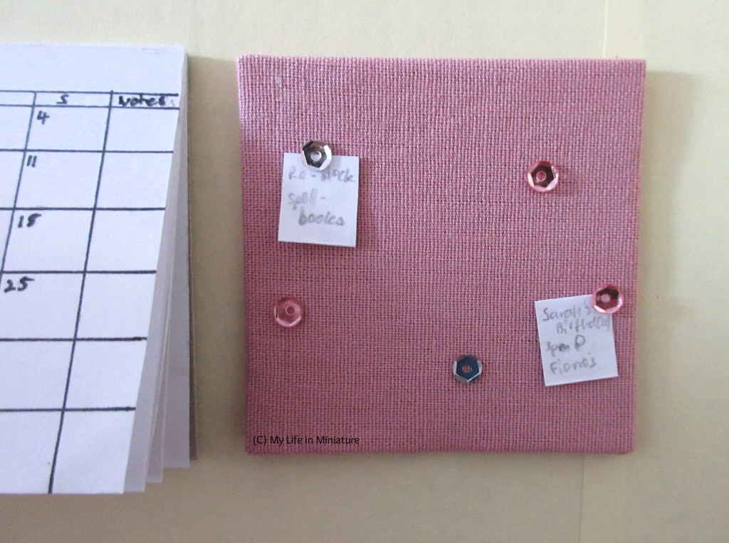 Close-up of the small noticeboard. It is covered in pink fabric, and has a few grey and pink pins in it. Two pins hold notices - one says 'Re-stock spell-books', the other says 'Sarah's Birthday! 3pm @ Fierro's'. 